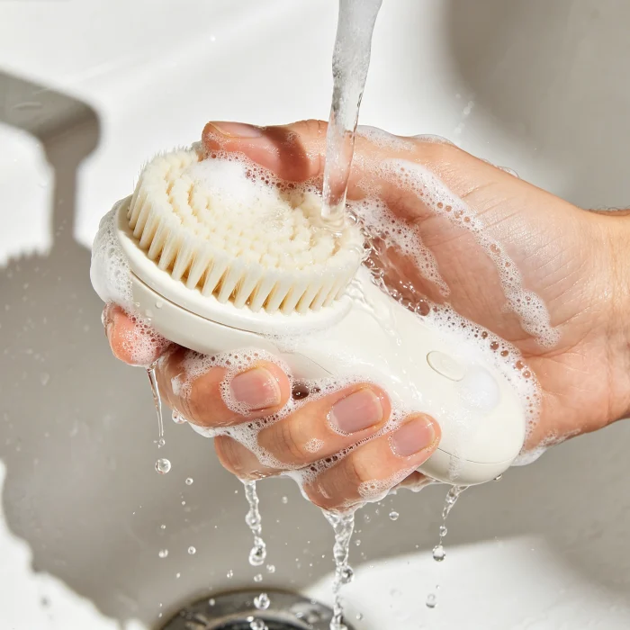 a person washing a facial cleansing brush under running water with soap suds